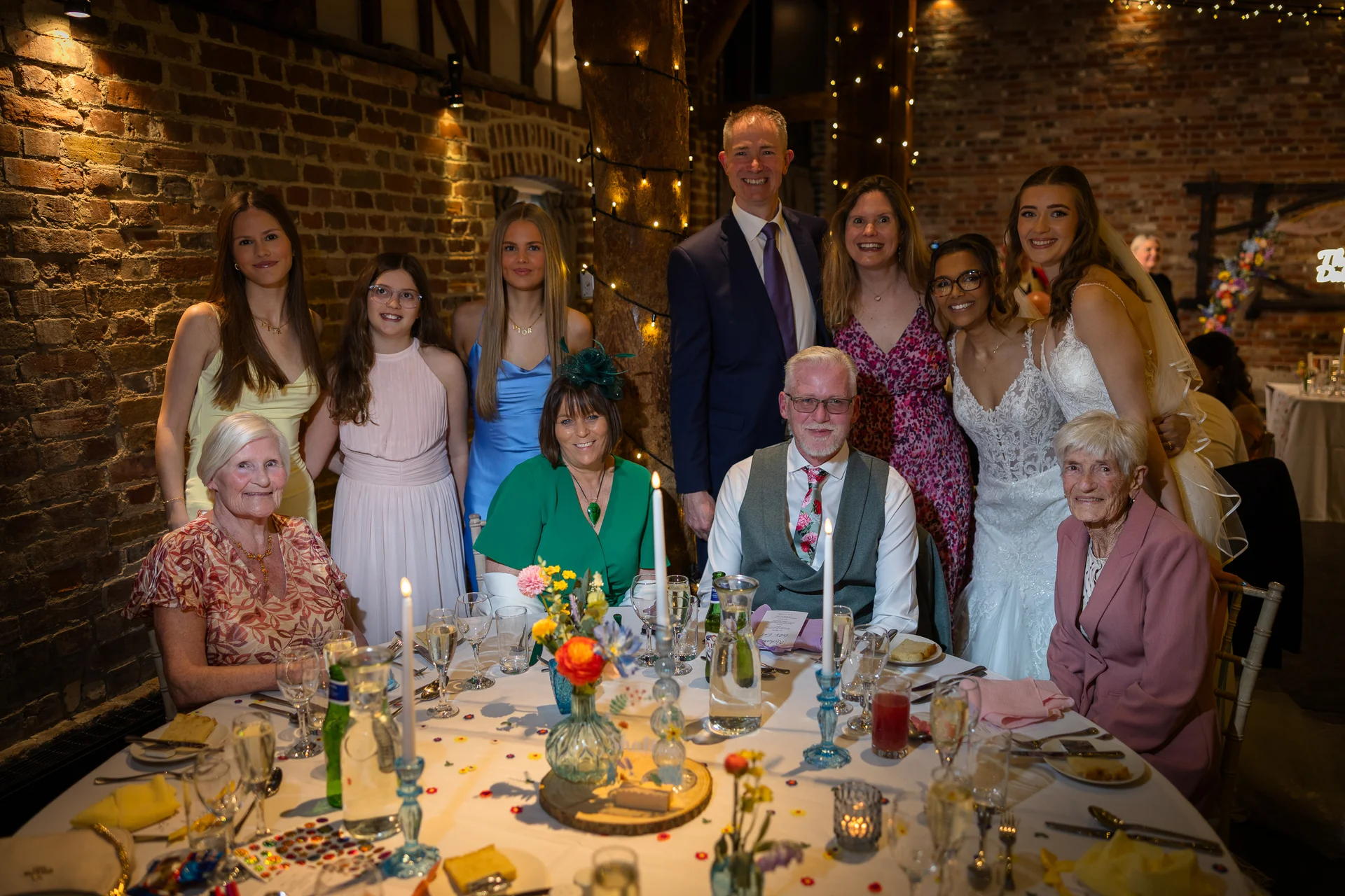 A large wedding group smiles warmly around a candlelit round banquet table inside an exposed brick barn decorated with fairy lights.