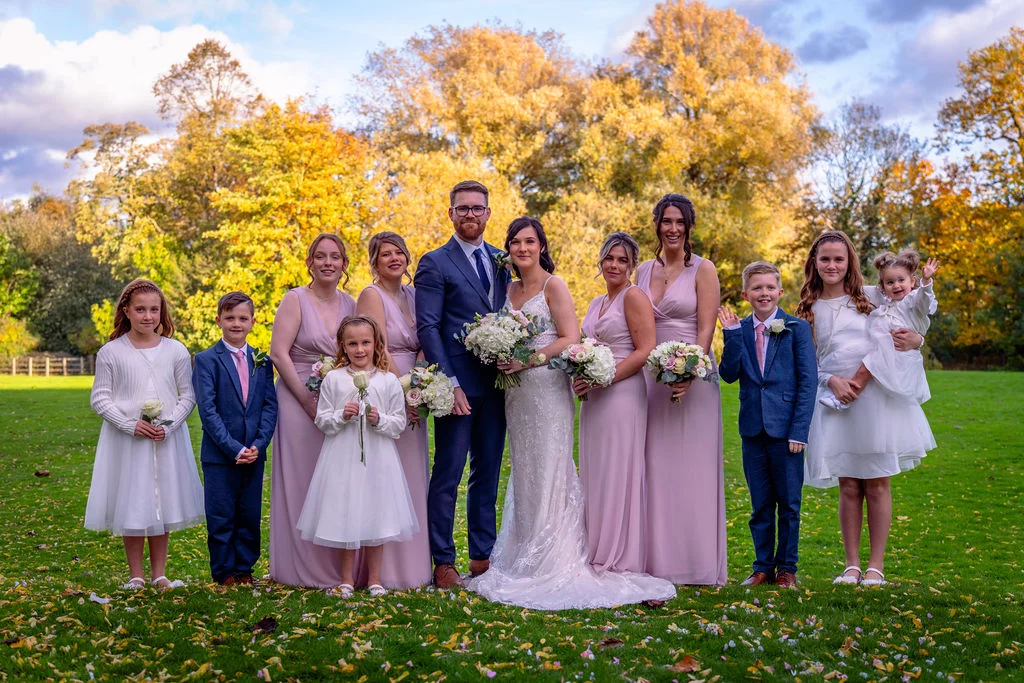 A bridal party poses outdoors on a grassy lawn surrounded by vibrant autumn trees under a partly cloudy sky.