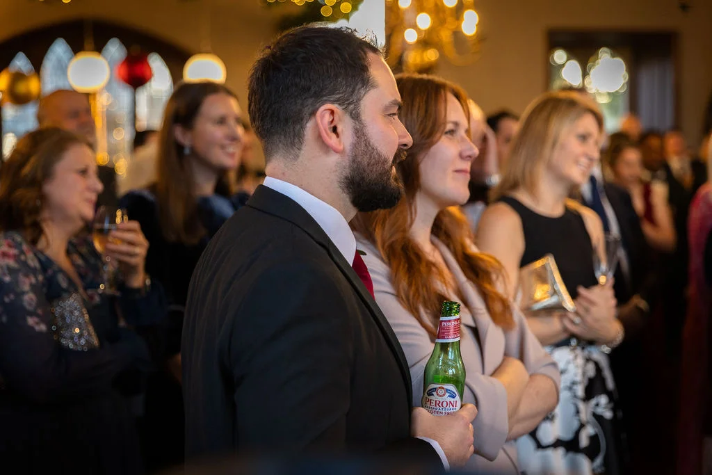 A group of wedding guests smiling and watching events unfold indoors, with a bearded man in a suit holding a bottled drink in the foreground.