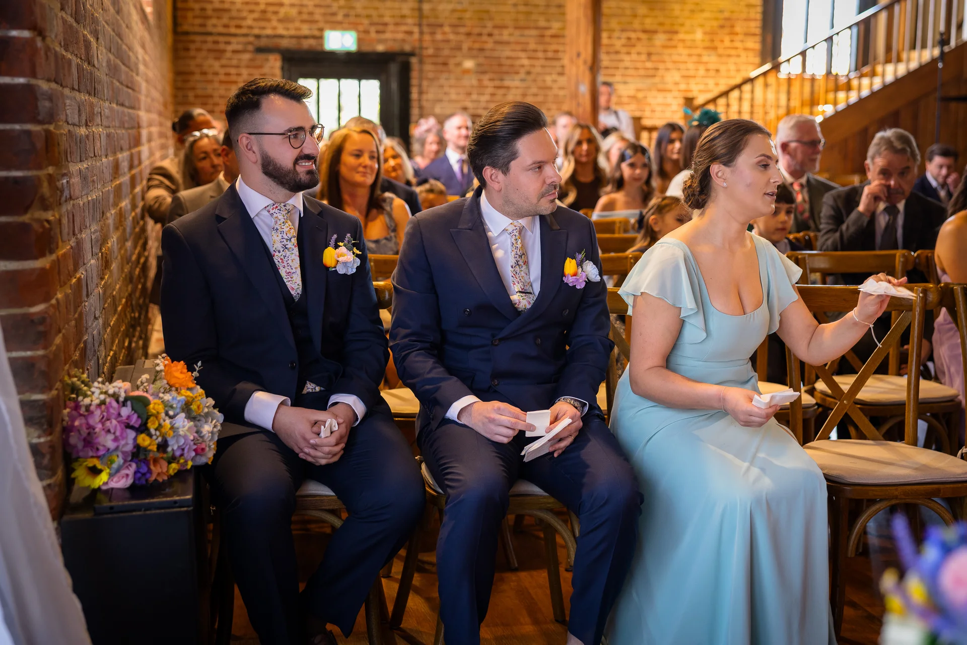 A group of wedding guests seated on wooden chairs inside an exposed brick barn venue, with two men in navy suits wearing floral ties and a woman in a sage green dress holding tissues.