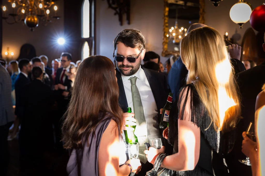 Wedding guests mingle and chat during an evening reception in a historic venue illuminated by warm chandeliers and soft natural light.