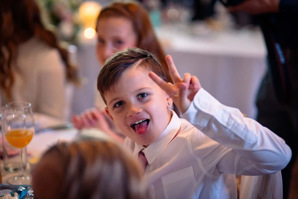 A young page boy wearing a white shirt makes a playful gesture with a peace sign and his tongue out while seated at a wedding reception table.