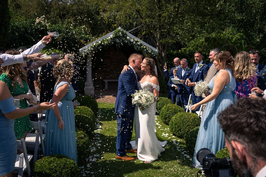Newlyweds share their first kiss as a married couple outdoors in a garden venue while guests shower them in confetti.