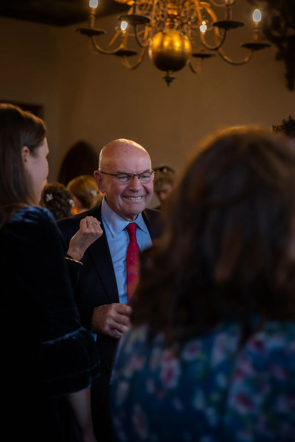 A smiling older male wedding guest in a suit and patterned red tie mingles with guests under a large chandelier.