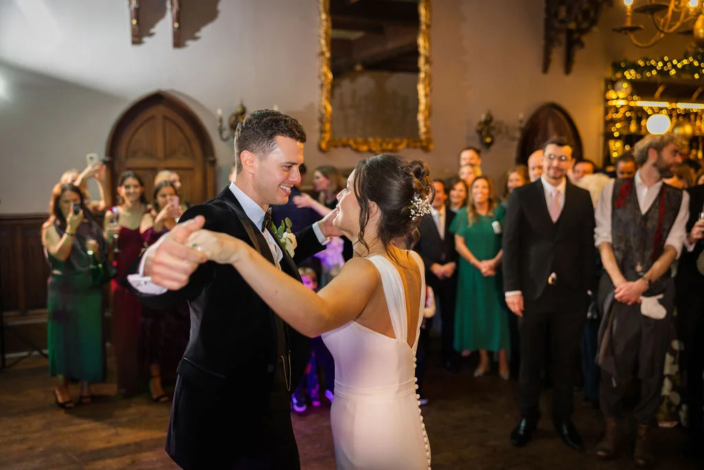 A newlywed couple shares their first dance while smiling lovingly at each other, surrounded by guests in a warmly lit, historic hall.