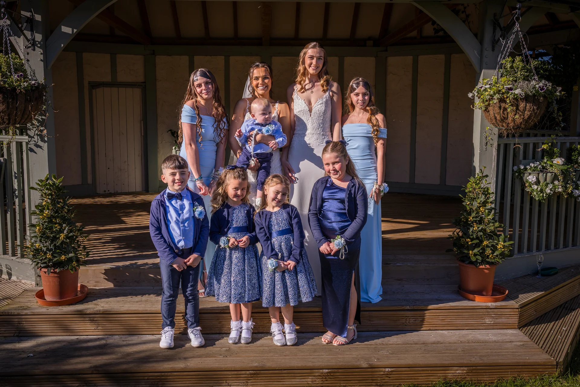 A bride in a lace wedding gown poses outdoors on wooden steps with her bridesmaids in pale blue dresses, alongside young flower girls and page boys.