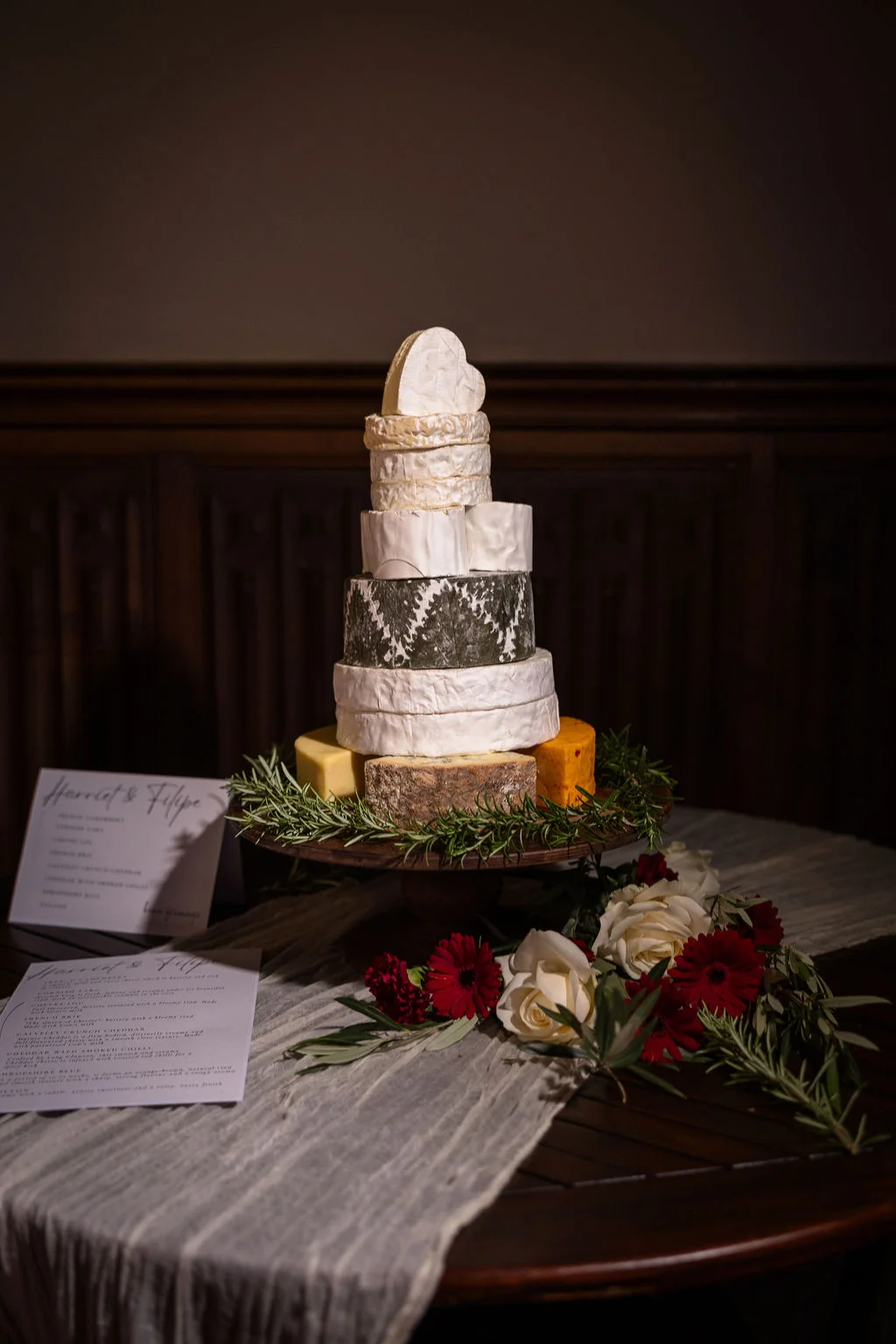 A multi-tiered wedding cheese tower stands on a wooden cake stand adorned with rosemary, white roses, and dark red flowers, accompanied by printed menus on a textured table runner.