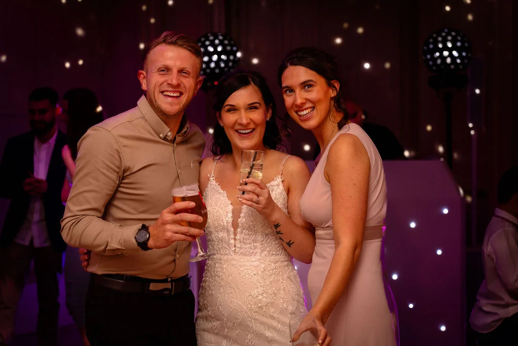 A smiling bride holding a drink stands between a guest with a beer and another guest during the evening reception.