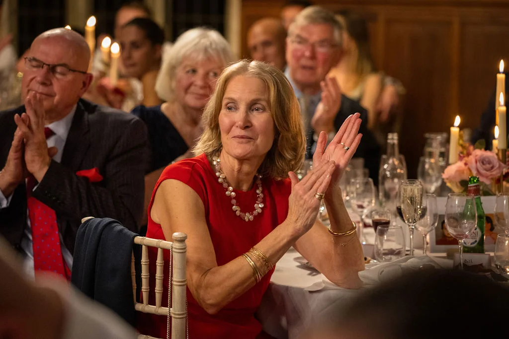 A smiling guest wearing a red dress and pearl necklace claps happily during wedding speeches at a candlelit reception.