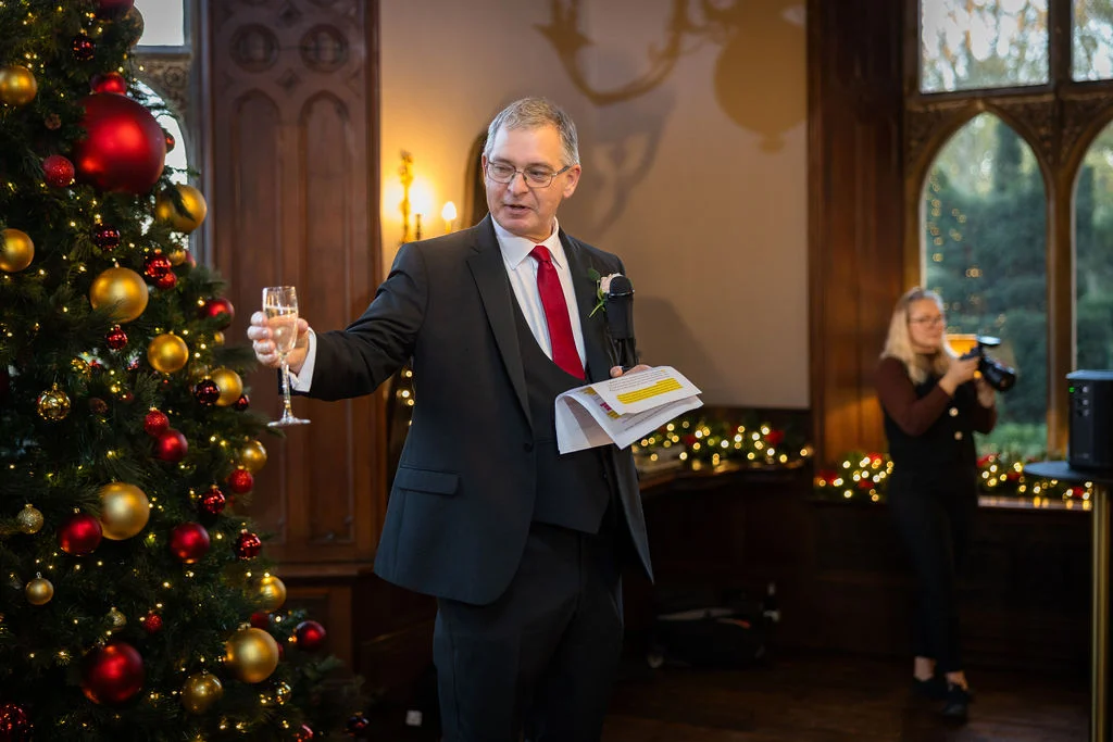 A smiling man in a dark three-piece suit delivers a wedding speech next to a large, festively decorated Christmas tree while holding a glass of champagne.