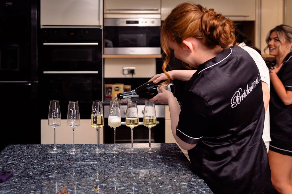 A bridesmaid in a black satin pyjama top pours champagne into personalised flutes lined up on a granite kitchen island during wedding morning preparations.