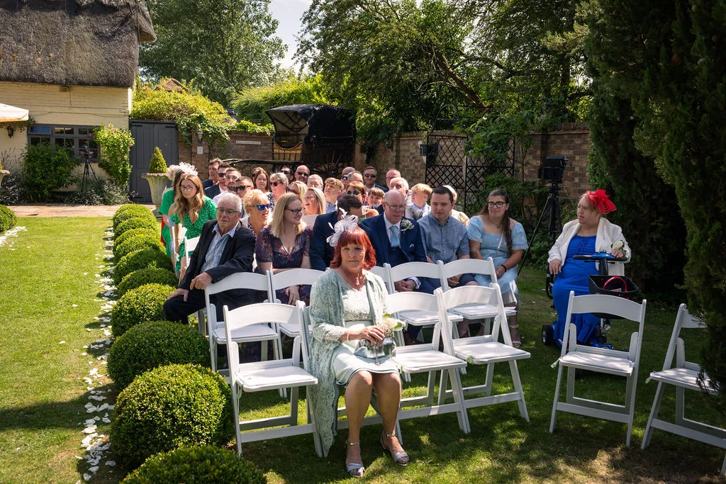 Wedding guests seated on white chairs on a manicured lawn during an outdoor ceremony in a picturesque country garden.