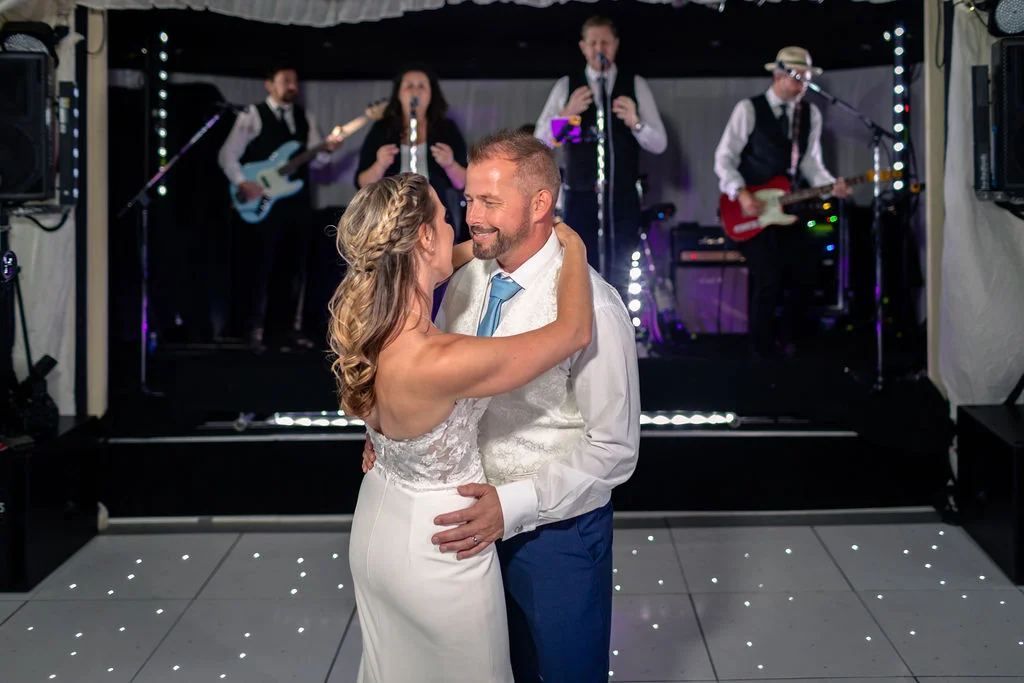 A newlywed couple shares their romantic first dance on a lighted dance floor while a live band performs on stage behind them.