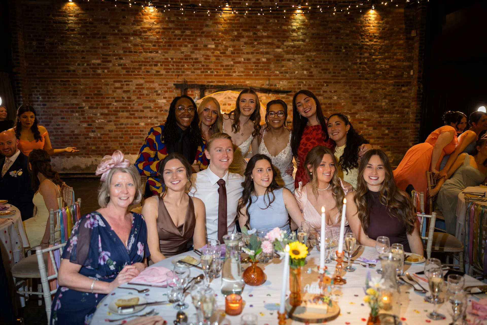 A large group of wedding guests smiling warmly together around a candlelit banquet table inside a rustic venue with exposed brick walls.