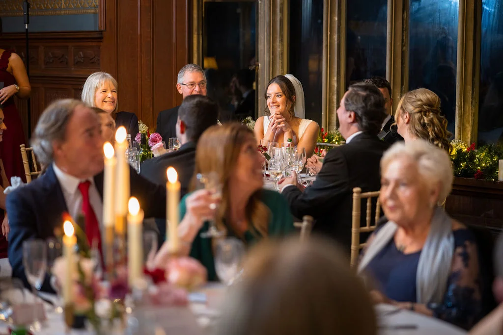A smiling bride listens attentively during wedding speeches at a candlelit reception surrounded by family and guests.