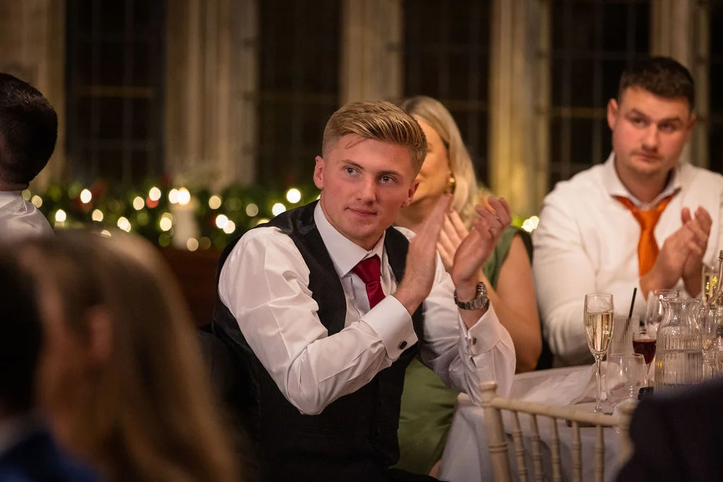 A guest smiles and applauds during the wedding speeches, seated at a candlelit reception table with champagne glasses.