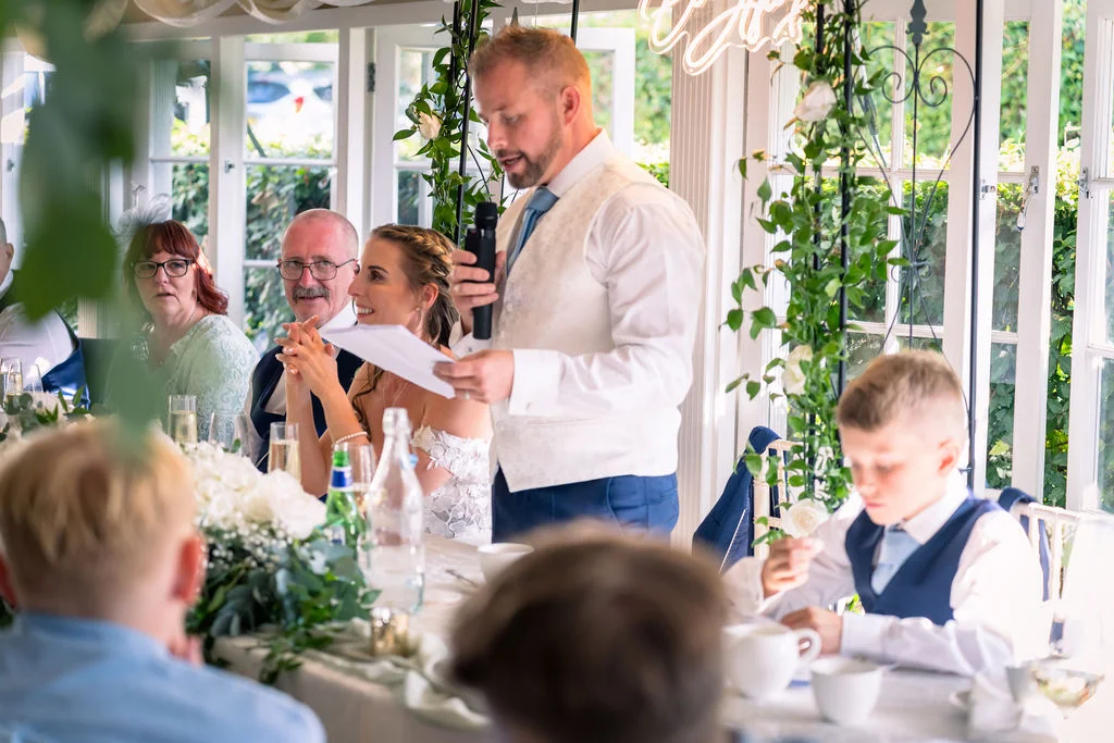 A groom delivers a speech reading from notes while holding a microphone at the top table in a light-filled orangery wedding venue.