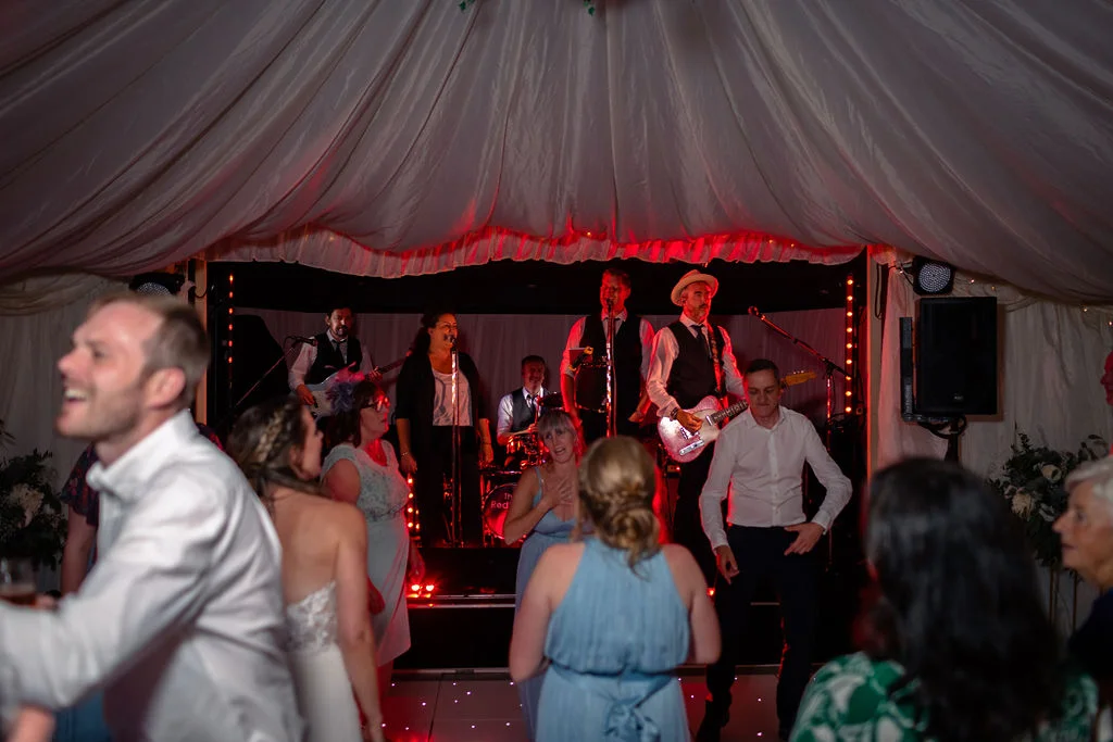 Wedding guests enjoy the evening reception dancing in front of a live band performing on a stage inside a marquee draped with white fabric.
