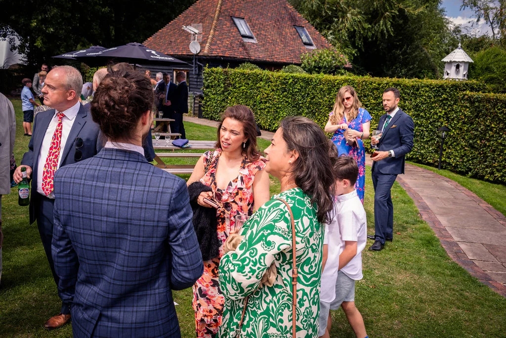 Wedding guests mingle and chat outdoors on a sunny day during the drinks reception in a manicured garden setting.