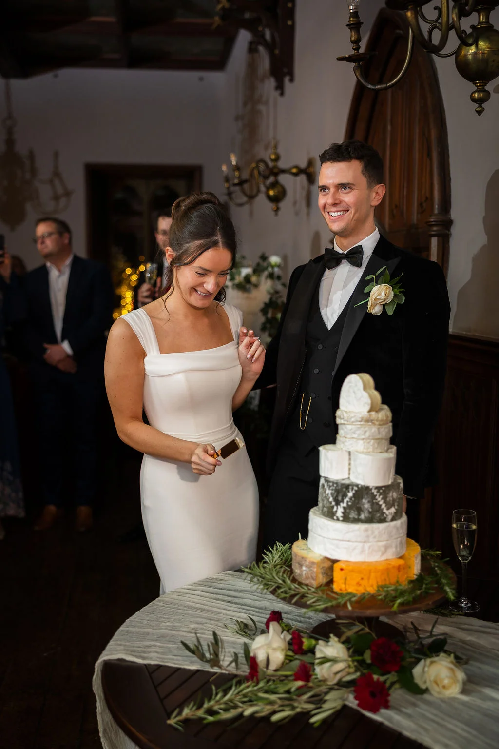 A smiling bride and groom cut a unique tiered cheese wedding tower decorated with rosemary and white roses during their evening reception.