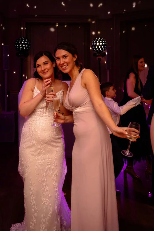 A smiling bride in a lace wedding dress and her bridesmaid holding champagne glasses pose together at an evening wedding reception with disco lighting.