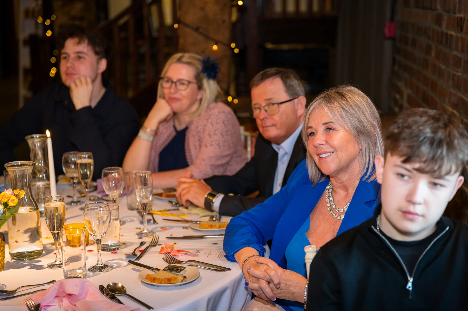 Wedding guests seated along a long banquet table during speeches, smiling and listening attentively in a rustic venue with exposed brick walls.