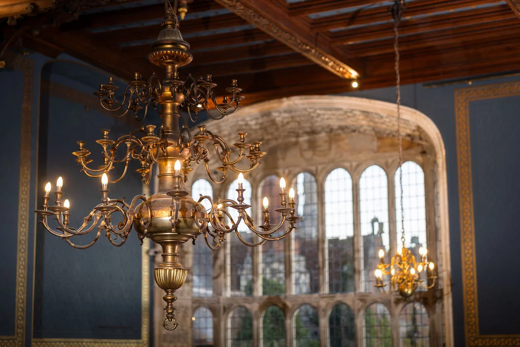 An ornate brass chandelier hangs in the foreground of a grand historic venue with a vaulted wooden ceiling, stone archways, and large windows.