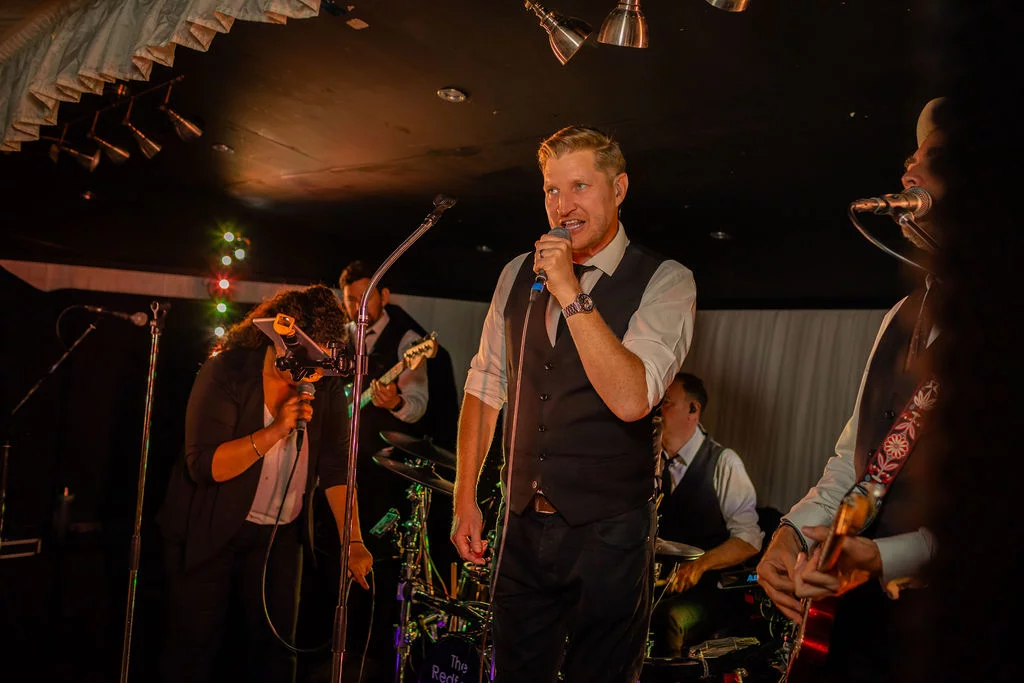 A live wedding band performs on stage during the evening reception, with a male vocalist singing energetically into a microphone.