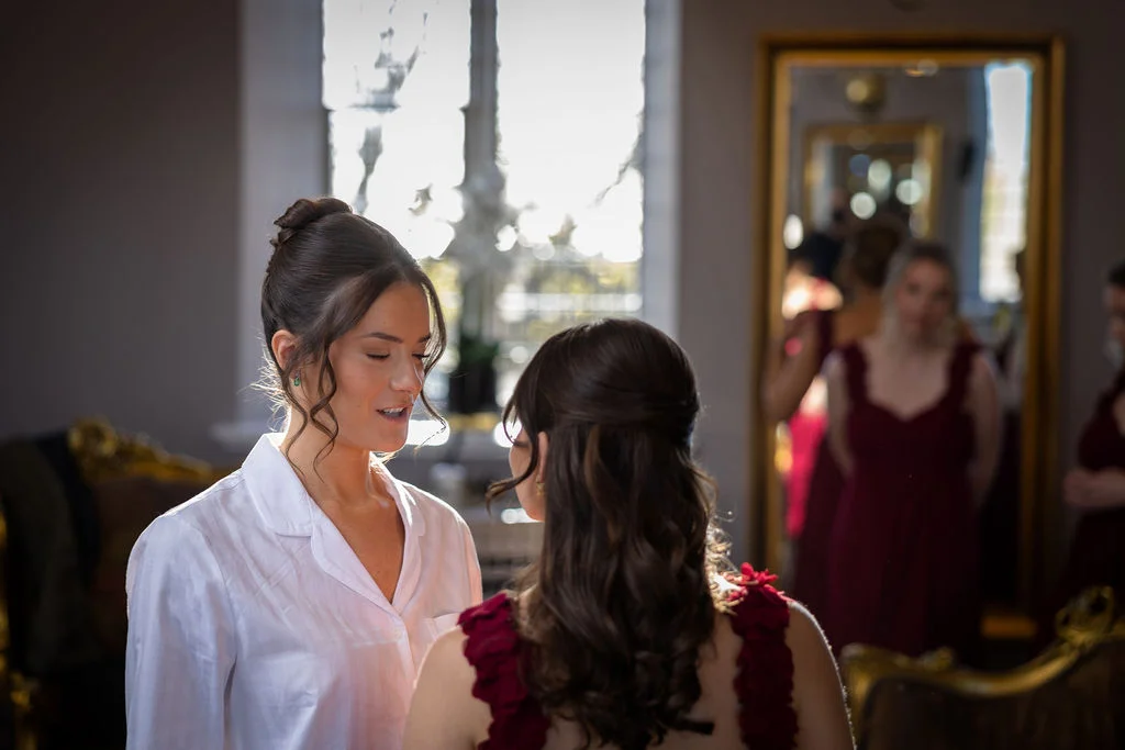 A bride in a white button-down shirt shares a quiet, tender morning moment with her bridesmaid wearing a burgundy dress in a sunlit room.