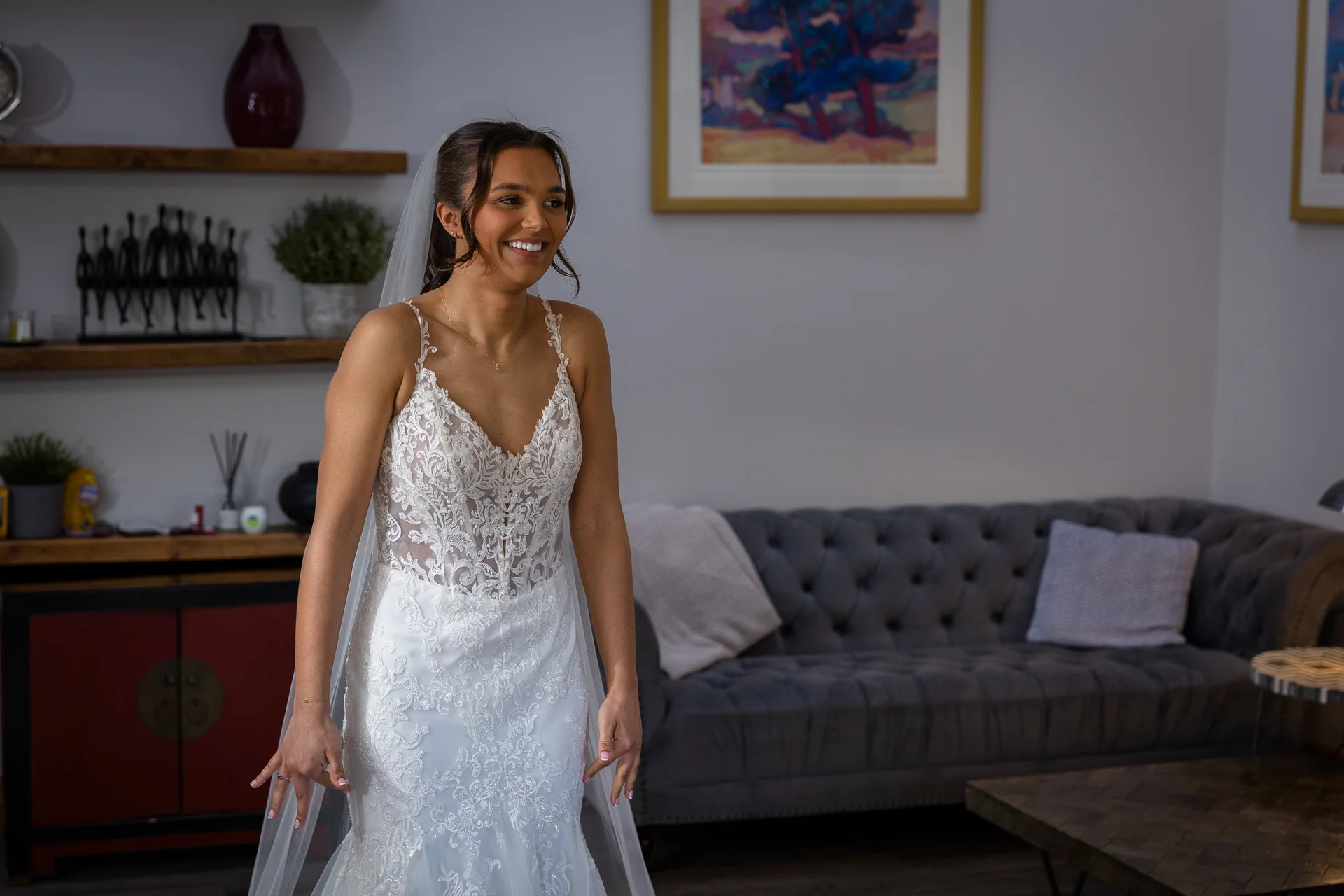 A smiling bride wearing a lace wedding dress with a sheer corset bodice and v-neckline stands indoors during bridal preparations.