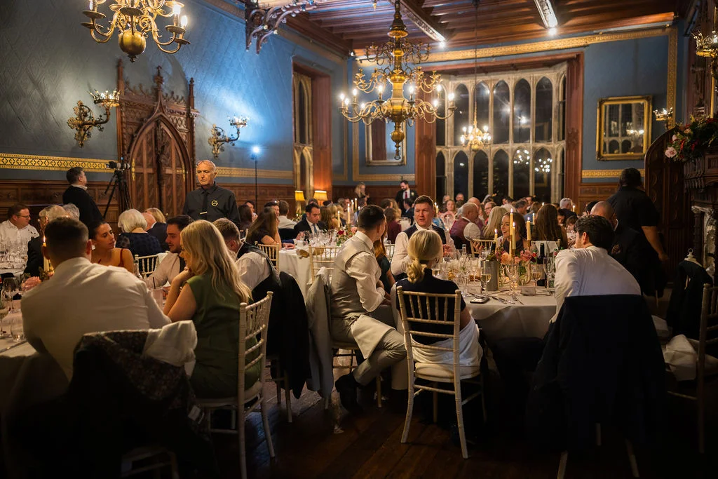Guests enjoy a wedding breakfast inside a grand, panelled ballroom featuring magnificent chandeliers and large arched windows illuminated by warm candlelight.