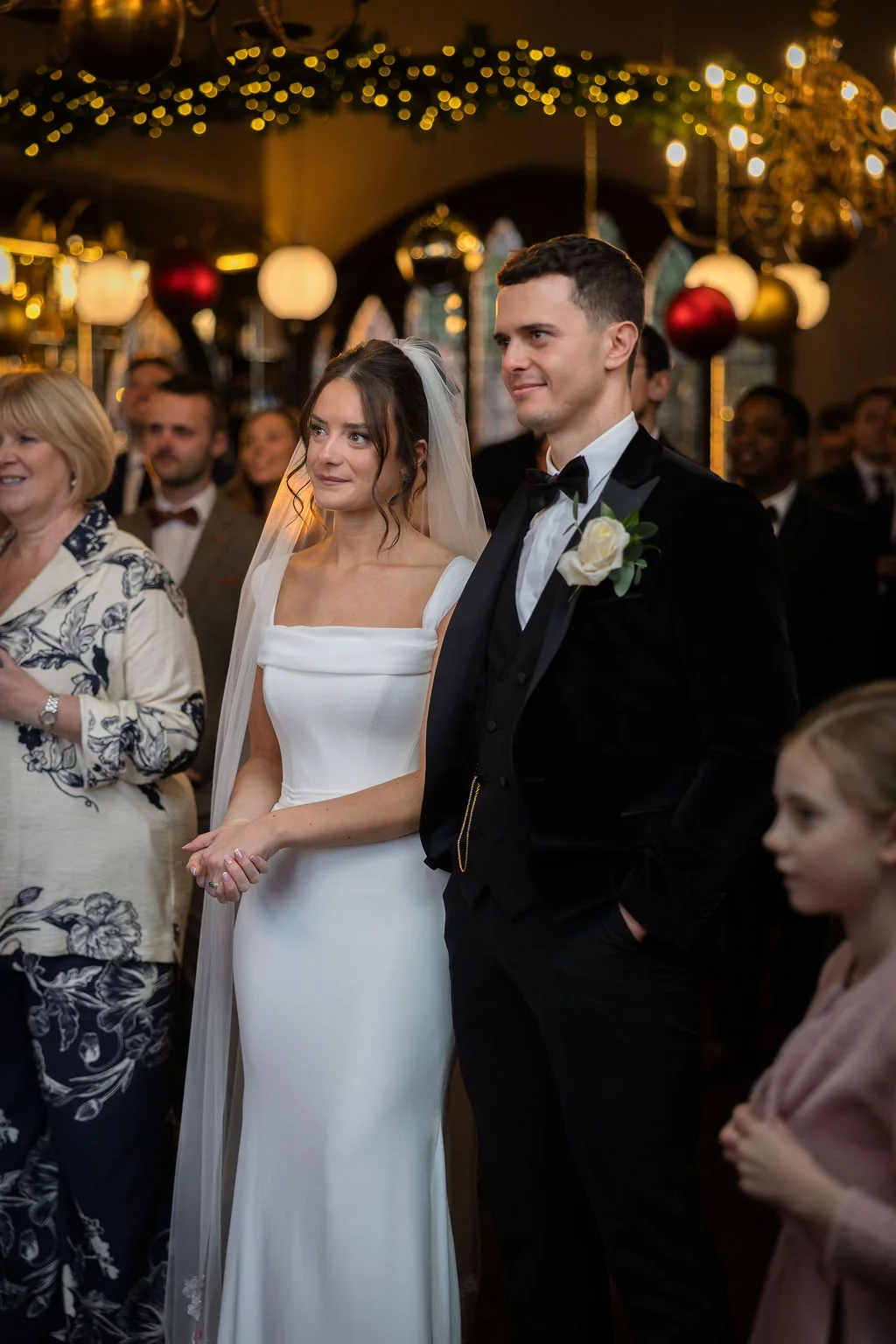 A newlywed bride in a sleek square-neck gown and veil stands beside her groom in a black velvet tuxedo during a candlelit winter wedding ceremony.