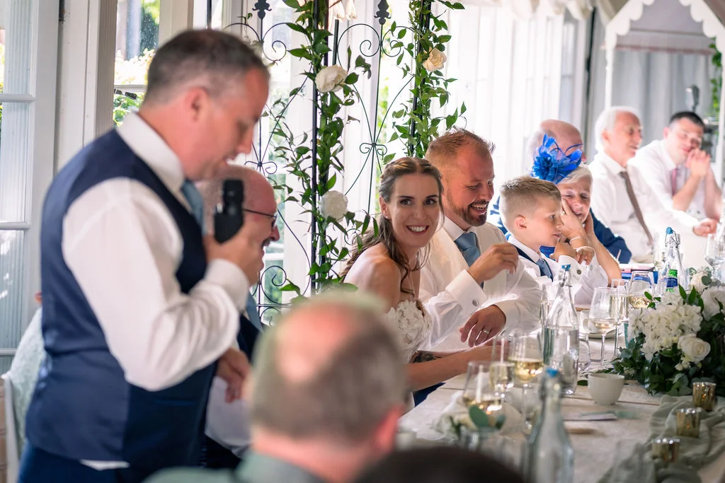 A wedding guest stands to deliver a speech at the top table while the bride, groom, and other guests laugh and listen attentively.
