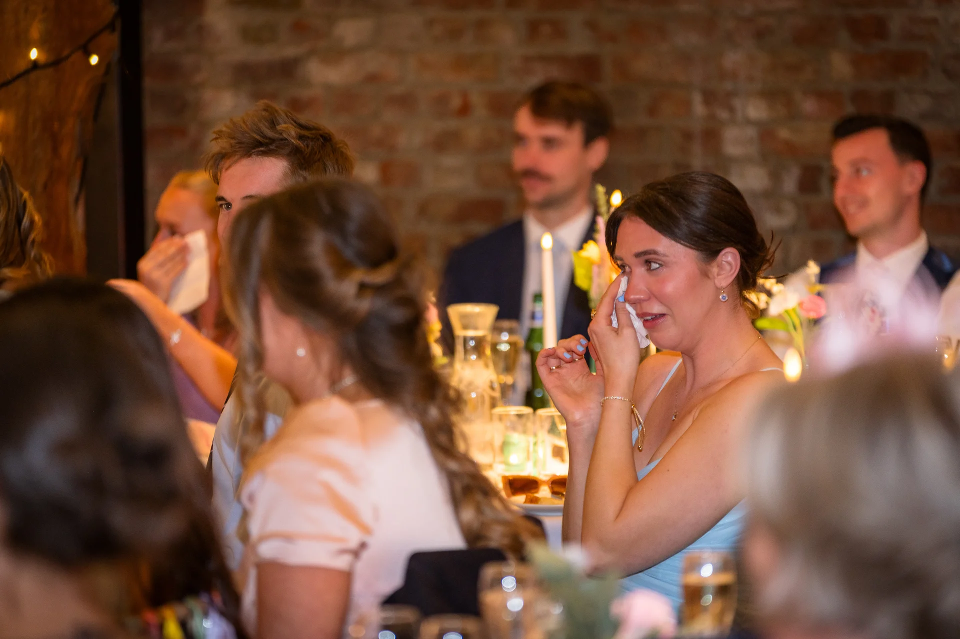 An emotional wedding guest wiping away tears with a tissue during the wedding speeches inside a venue with exposed brick walls.