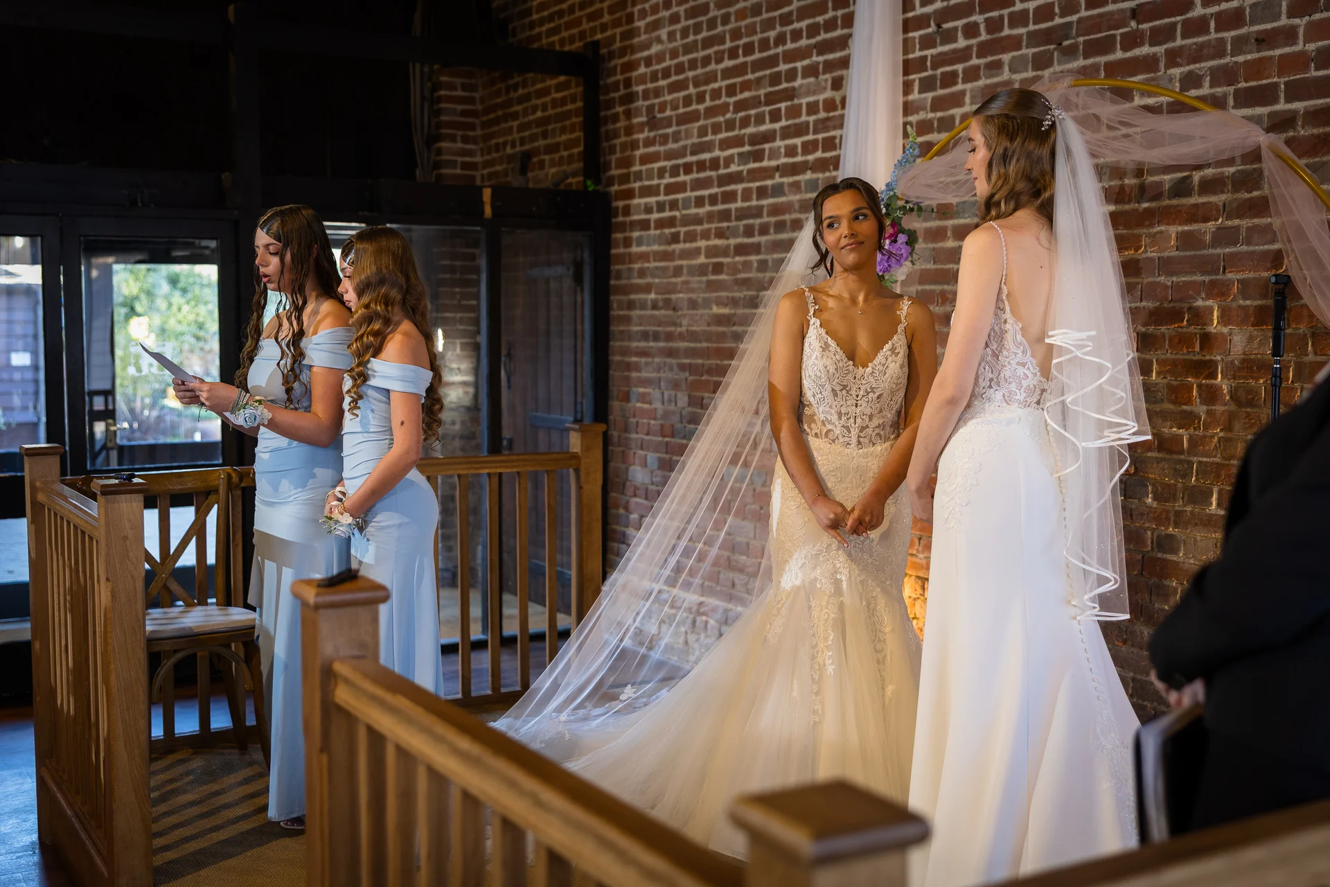 Two brides stand together at the altar during a barn wedding ceremony, accompanied by bridesmaids wearing pale blue dresses against an exposed brick backdrop.