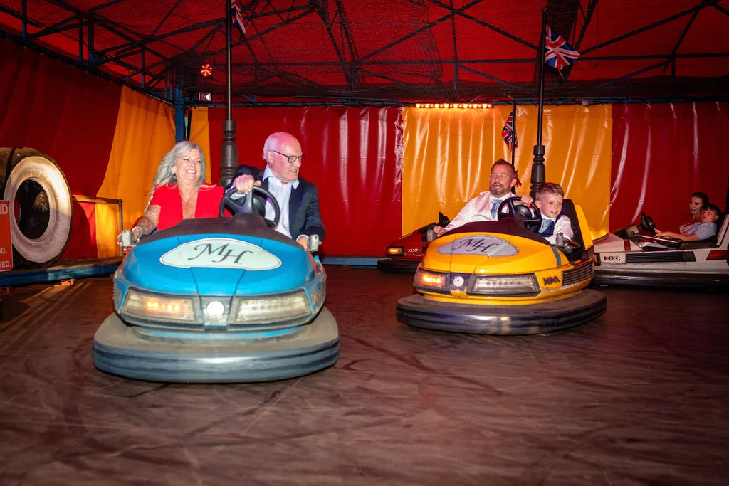 Wedding guests laughing and having fun riding bumper cars at a retro funfair attraction during the evening reception.