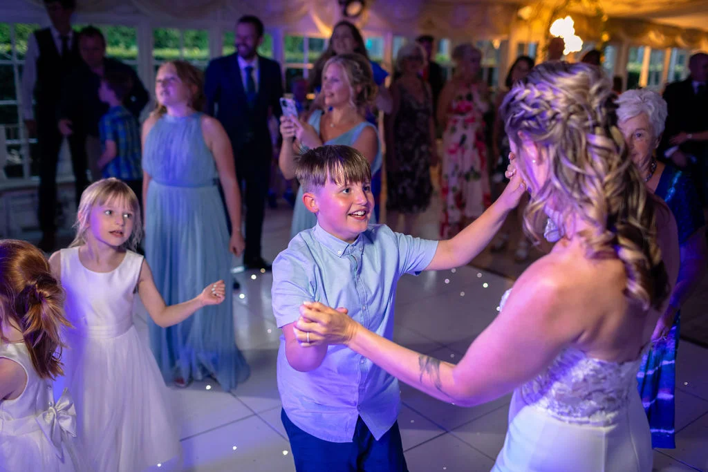 A young boy smiling brightly while dancing with the bride on a lit-up dance floor surrounded by wedding guests.