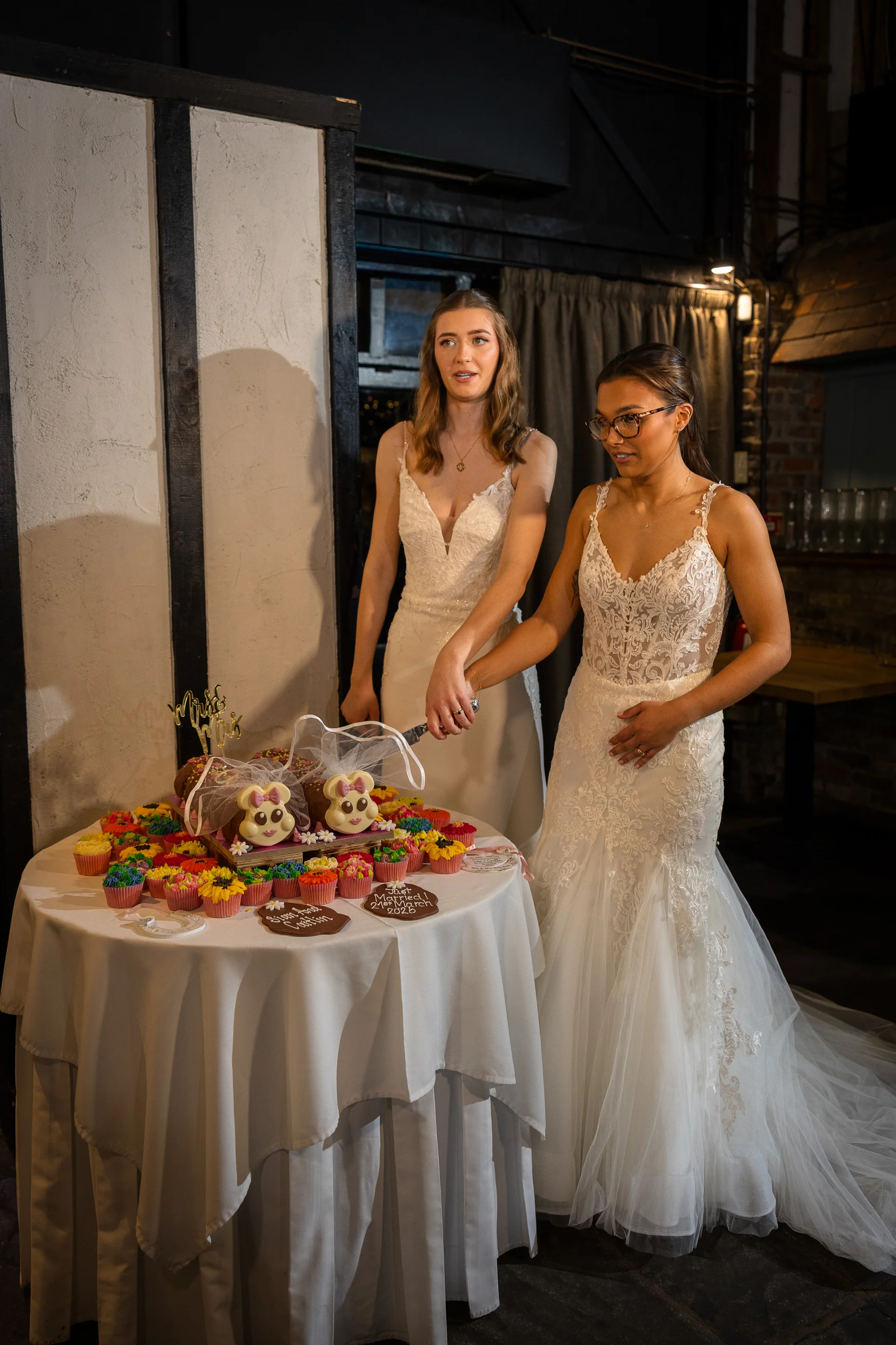 Two smiling brides in matching lace wedding gowns stand together to cut a whimsical cupcake tower with novelty animal toppers at their evening reception.