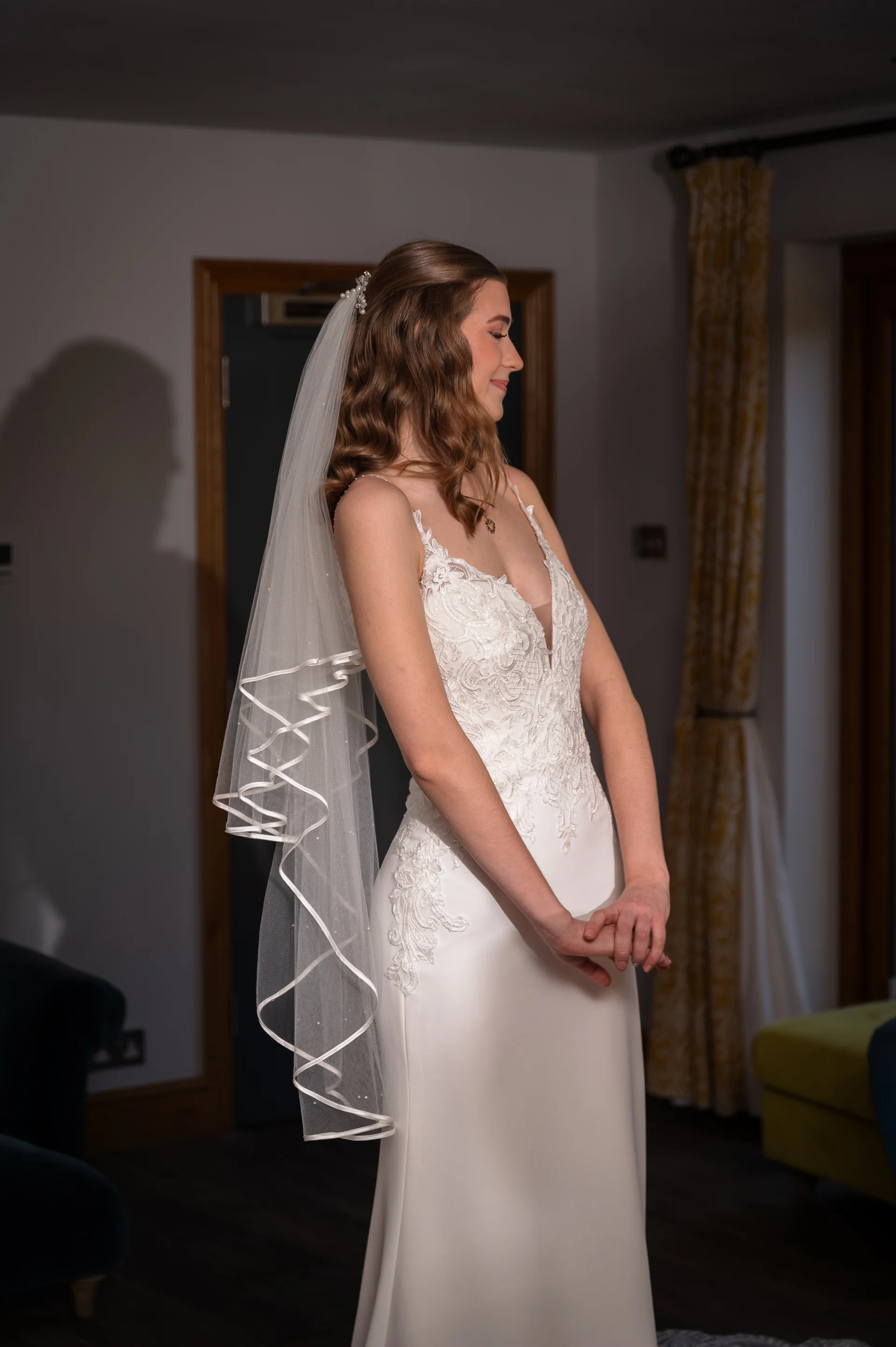 A side profile of a radiant bride in a V-neckline crepe dress with floral applique, wearing soft half-up waves and an elbow-length veil.