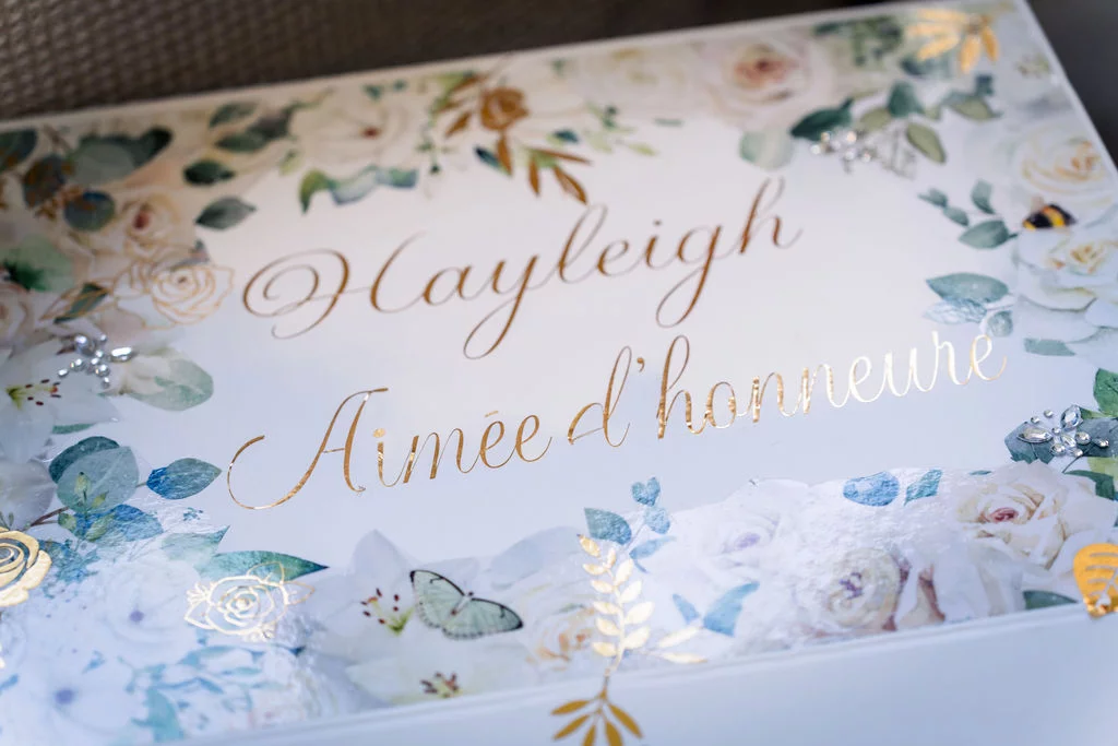 Close-up of a personalised white wedding box decorated with watercolour florals, butterflies, and elegant gold script reading 'Hayleigh' and 'Aimée d'honneur'.