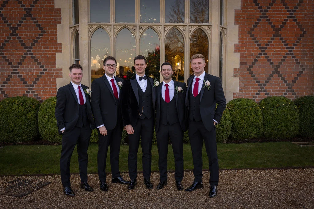 Five groomsmen in sharp black tuxedos and suits with red ties stand proudly outdoors in front of a grand manor house with large stained glass windows.