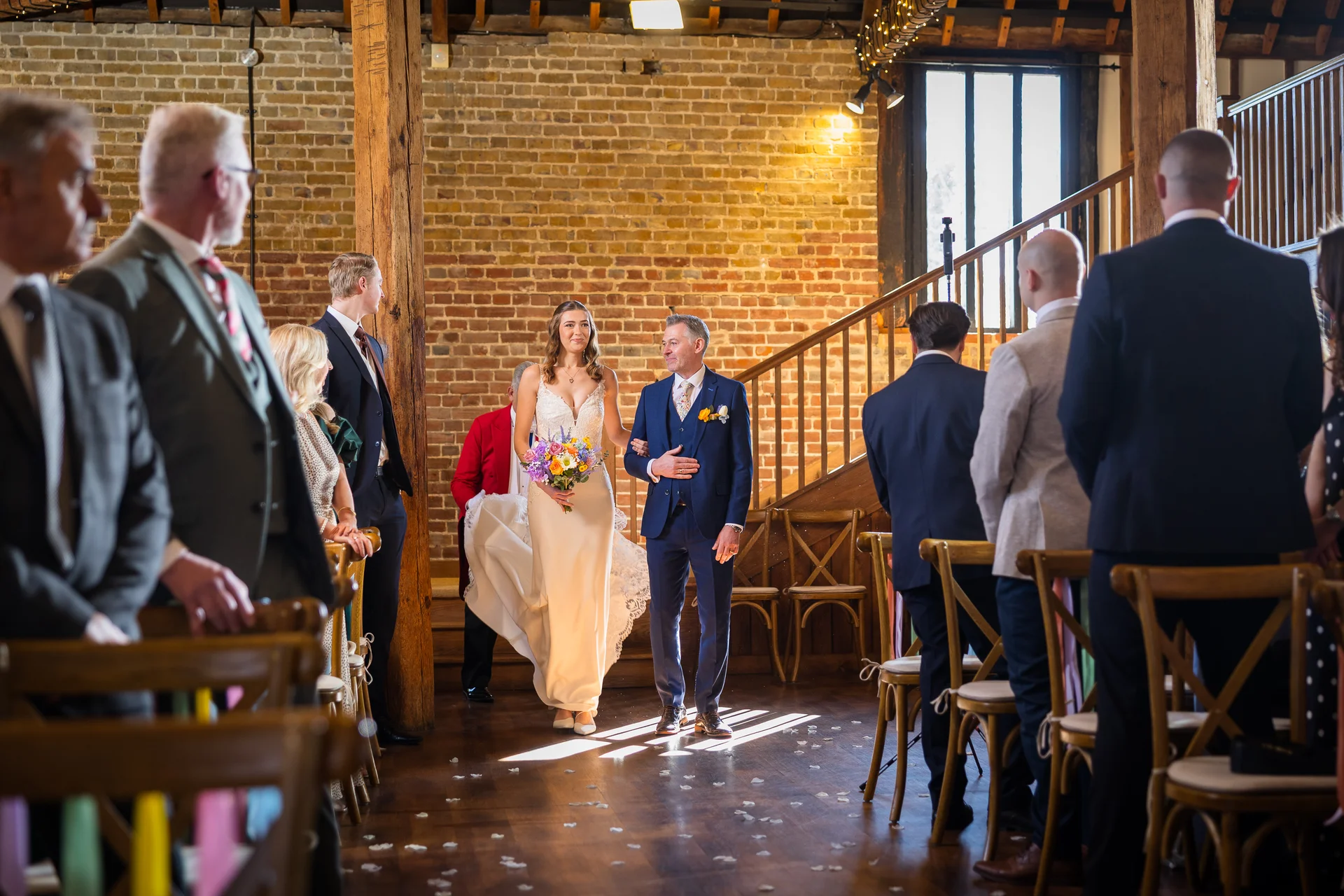 A smiling bride and groom walk down the aisle together inside a rustic brick barn wedding venue, watched by guests seated on wooden chairs.