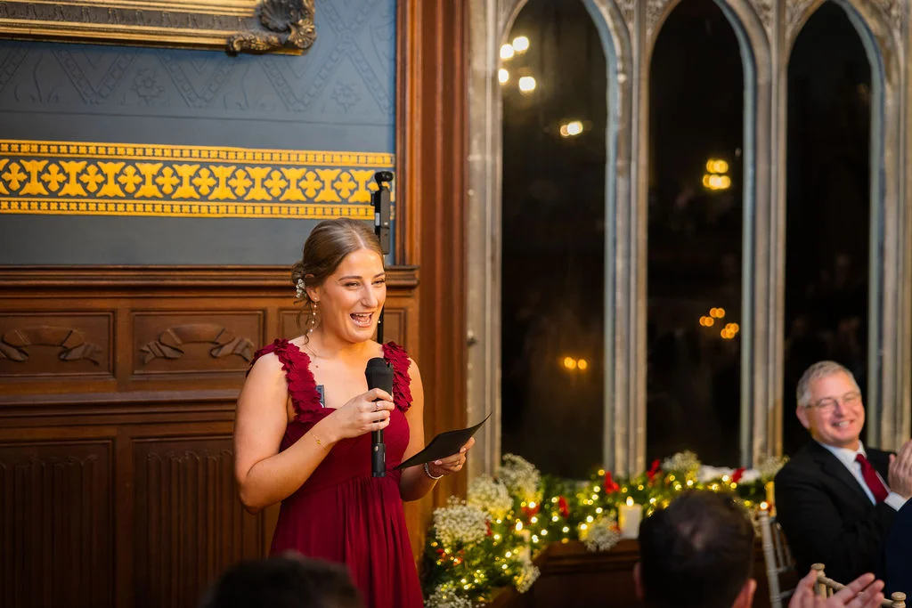 A bridesmaid wearing a burgundy dress delivers a speech with a microphone inside a wood-panelled hall decorated with glowing fairy lights.
