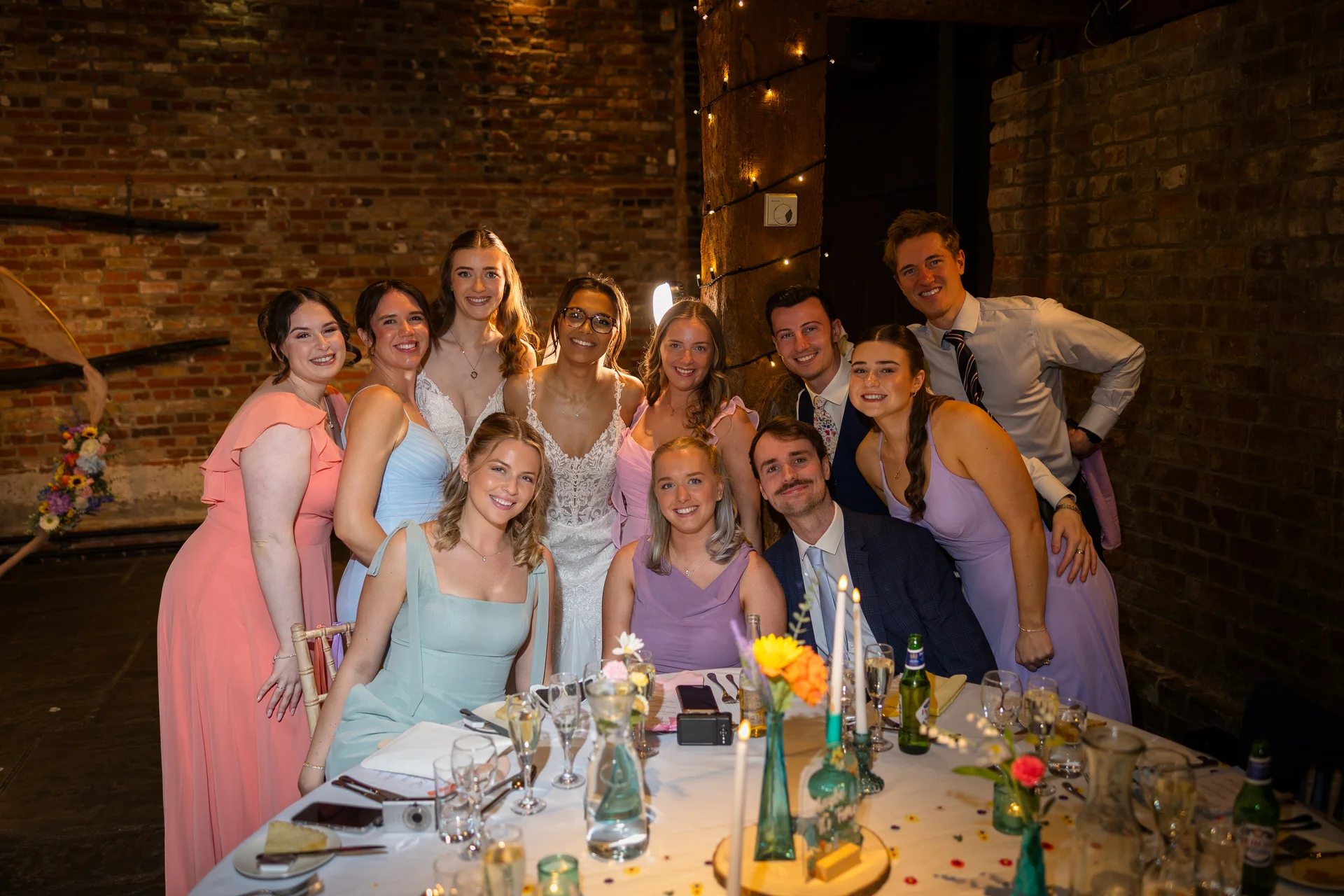 A happy group of wedding guests and bridesmaids gather around a beautifully decorated reception table inside a rustic brick barn lit by string lights.