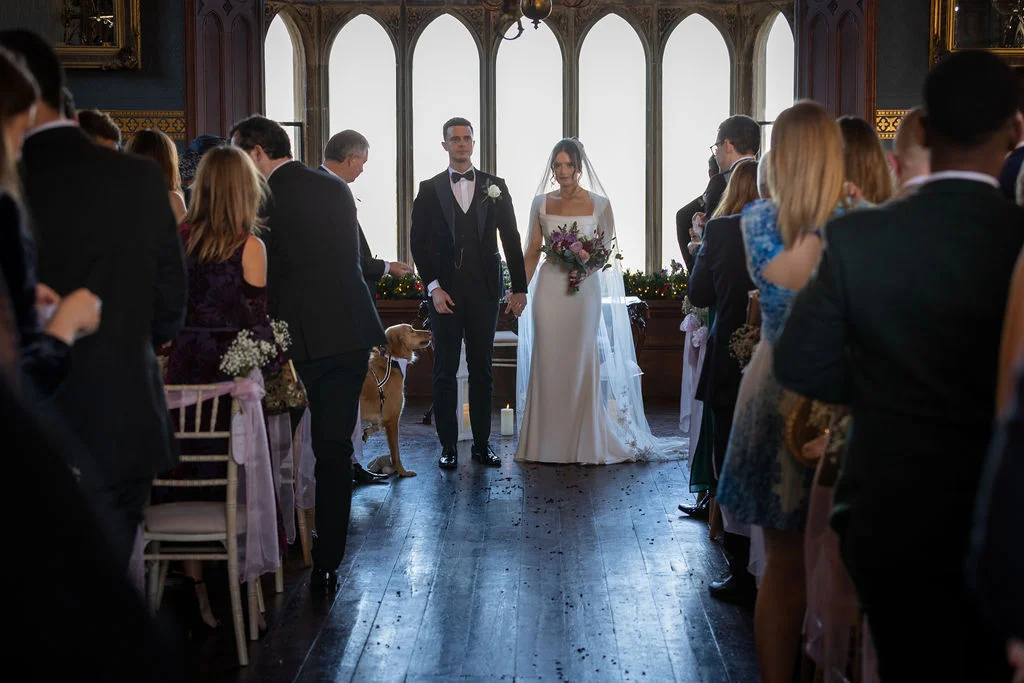 Newlywed bride and groom stand holding hands at the end of the aisle in a historic manor house ceremony room, framed by guests and a dog seated nearby.