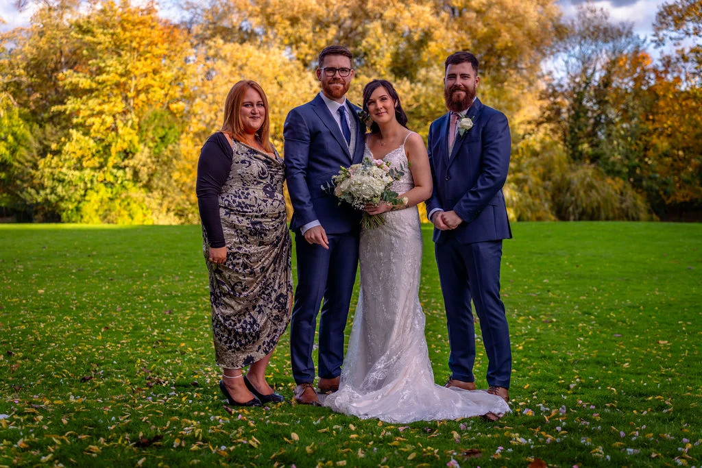 A bridal party poses outdoors on a vibrant green lawn scattered with fallen leaves, framed by golden autumn trees.