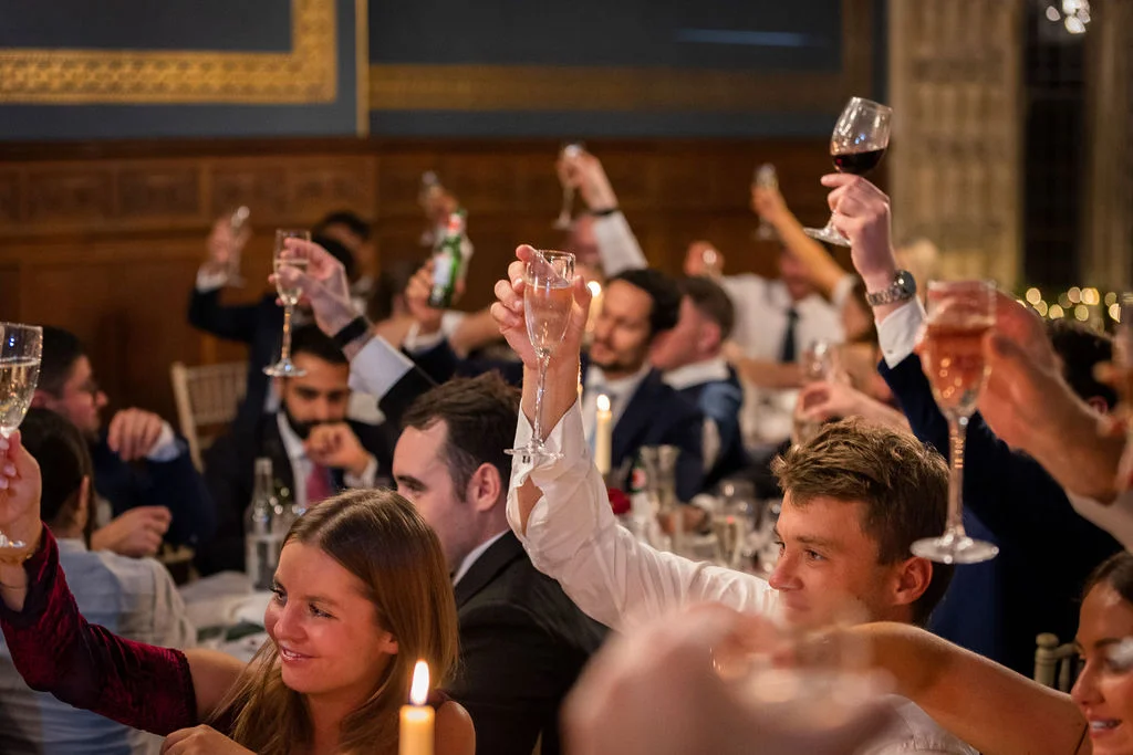 Wedding guests raise their glasses in a joyous toast during the reception speeches in a wood-panelled dining hall.