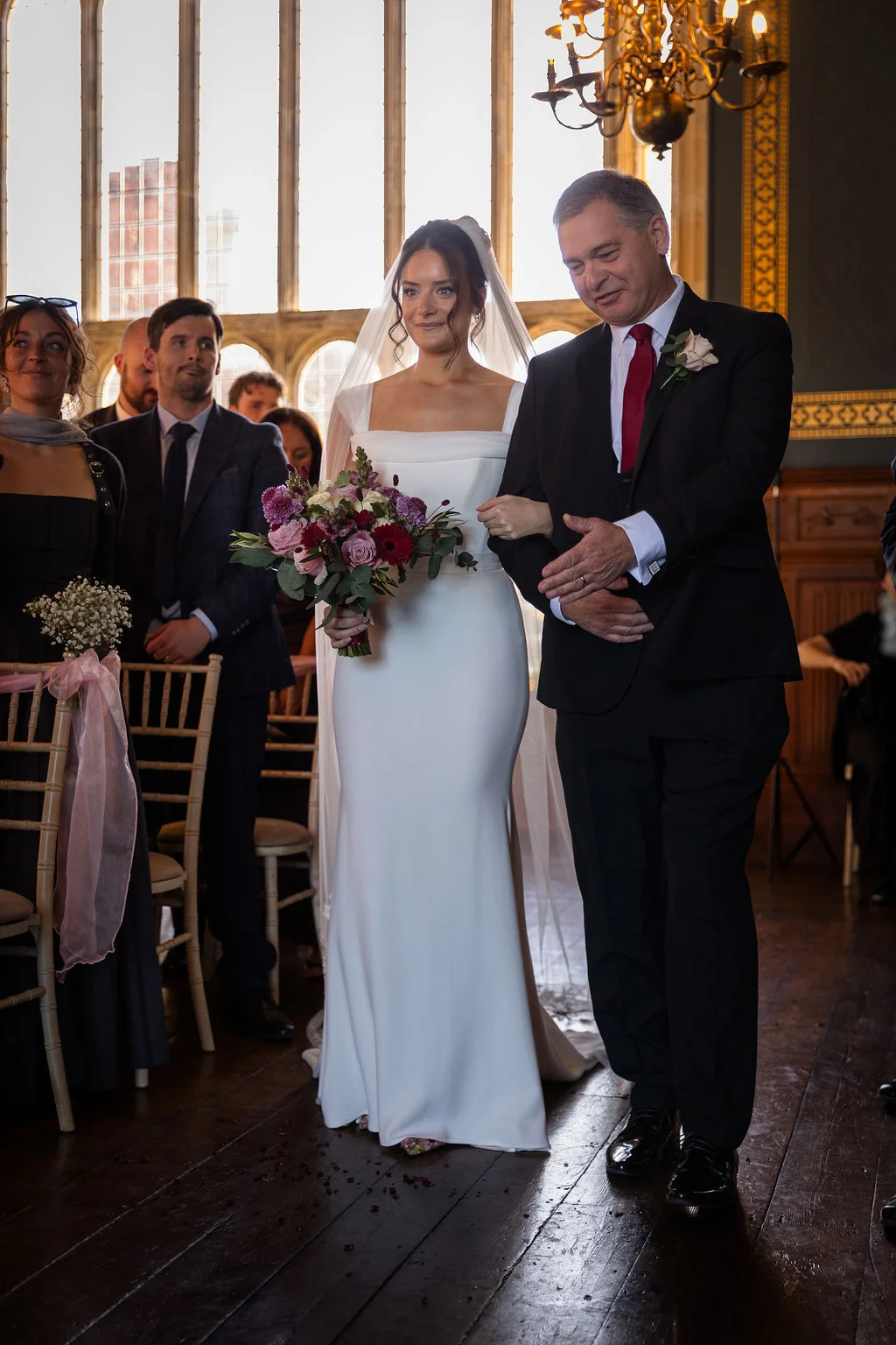 A smiling bride holding a colourful bouquet walks down the aisle arm in arm with her father inside a historic church with large windows.