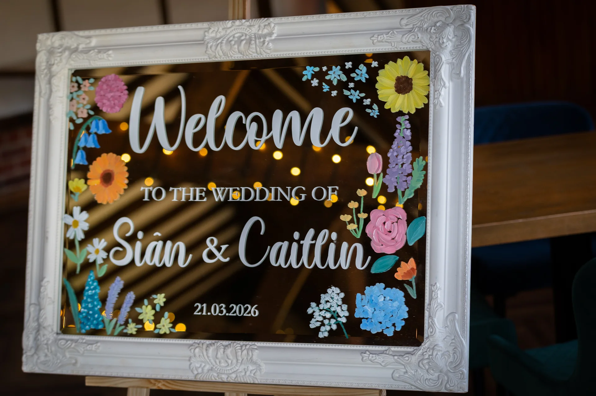 An ornate white framed mirror welcome sign on an easel displaying hand painted floral details and text welcoming guests to the wedding of Sian and Caitlin.