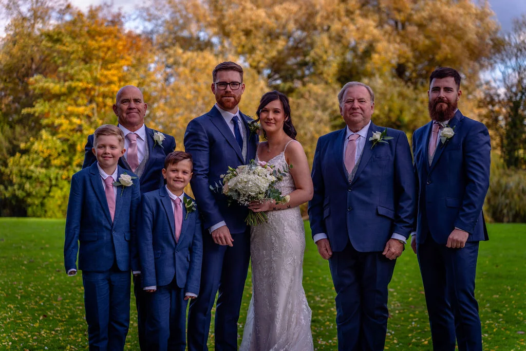 A bridal party including bride, groom, groomsmen, and young page boys pose together outdoors on a green lawn against a backdrop of golden autumn foliage.